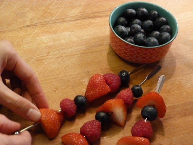 A child carefully threading a strawberry onto a skewer with rainbow fruit kebabs already in place