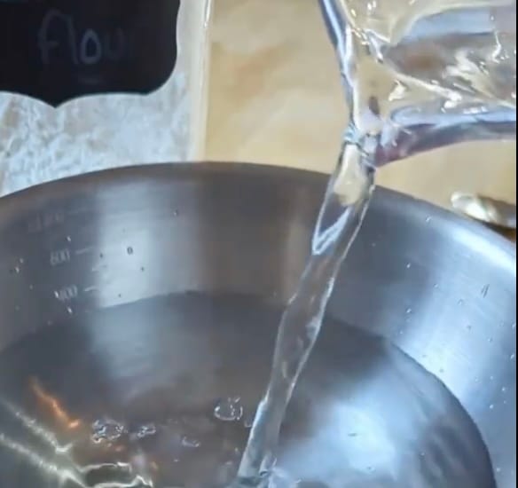 Pouring water from a small jug into a bowl on a kitchen worktop.  Pouring and transferring.